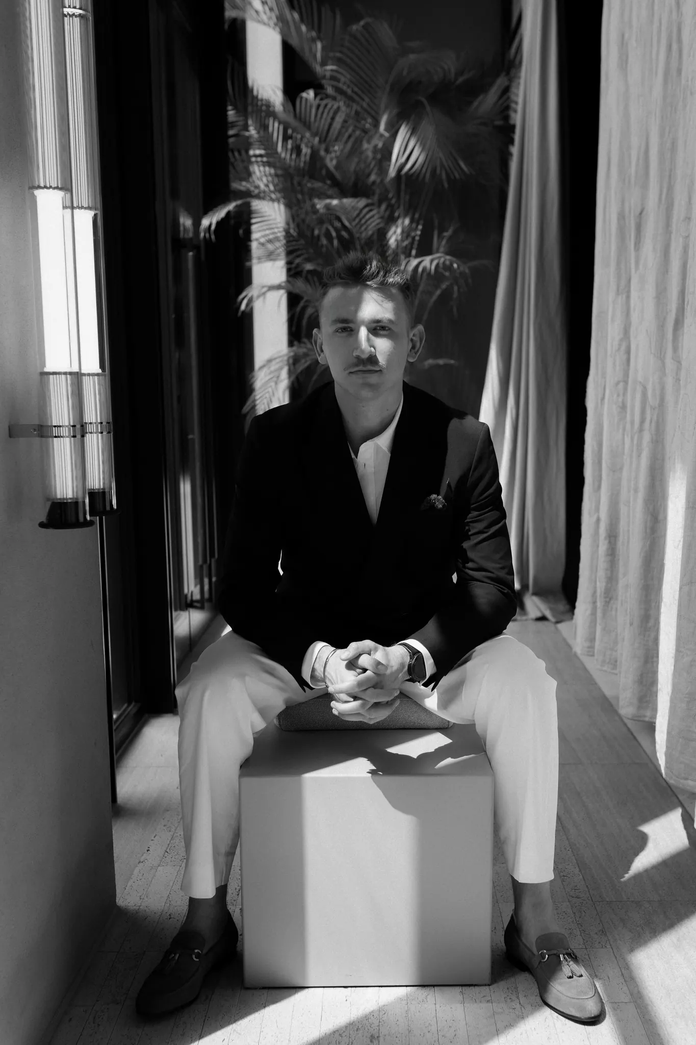 Sasha seated on a white cube beneath palm leaves, black and white