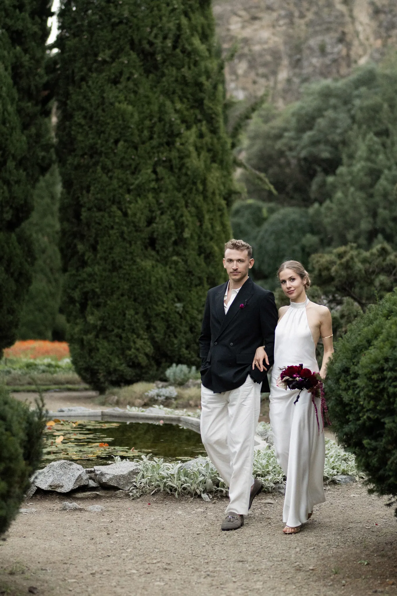 Veronika and Sasha walking past a lily pond, cypresses behind